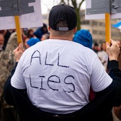 A photo of the back of a male protestor wearing a white T-shirt with "All Lies" written on the back. The man is wearing a black cap and is among a group of other protestors holding signs. We cannot see what is written on the signs are they are facing the other direction. 
