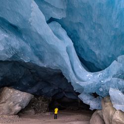 Person in yellow jacket stands inside a large ice cave with textured blue walls and rocky ground; Swiss Alps, Moteratsch Glacier