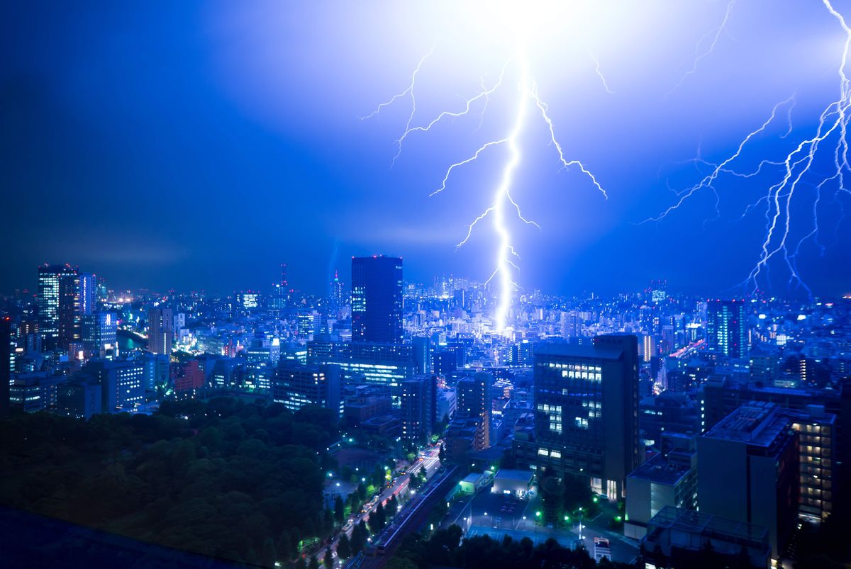 A photo taken from a high elevation at night shows Tokyo as a massive lightning strikes the ground in the middle of the image. The lightning is bright white, illuminating the city and casting a blue color around it. There are smaller, thinner lines of lightning to the right. 