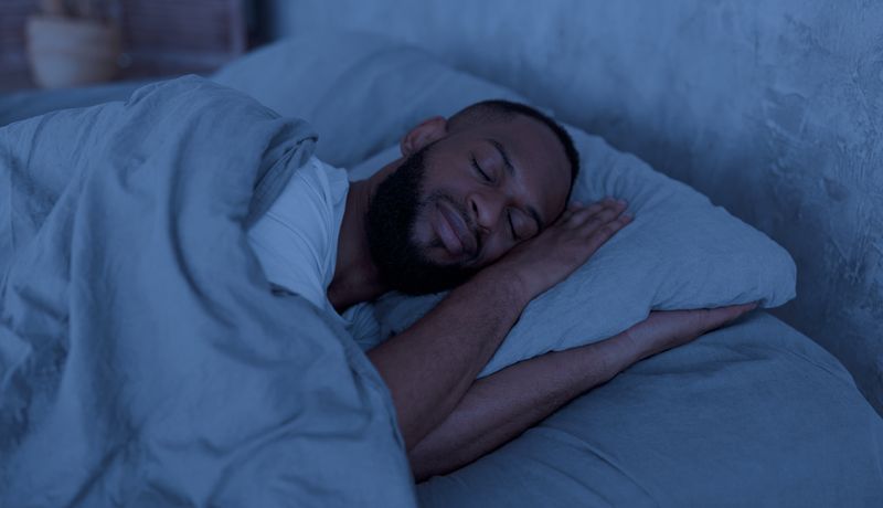 Portrait of happy young well-slept African American man lying in bed with closed eyes, resting in bedroom on the side in the dark night