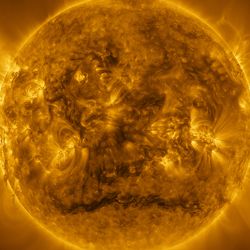 The Sun looks like a warm yellow sphere with a surface covered with glowing messy hair. The yellow glow extends to the edges of the image, with some regions brighter than others. Many bright yellow arcs stick out from a wide band around the Sun’s equator. A darker region stands out across a roughly horizontal line near the Sun’s south pole. The bright arcs and some darker material can also be seen around the Sun’s edges.